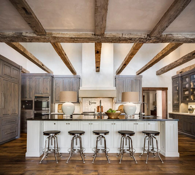 Rustic Kitchen. Rustic Kitchen kitchen features five Restoration Hardware Vintage Toledo Barstools positioned in front of a white kitchen island fitted with black marble countertops as well as two undermount sinks with satin nickel gooseneck faucets lit by white table lamps accented with beige shades. Under a stunning rustic ceiling beams a white plastered hood is located between stained pine kitchen cabinets mounted against a white glazed subway tile backsplash accenting white marble countertops. The room also features a stainless steel wall mounted double oven and a beautifully concealed pine paneled refrigerator located on an adjacent wall. The room is completed with glass front china cabinets located on the far right wall and rustic wood floors. #RusticKitchen Tiffany Farha Design