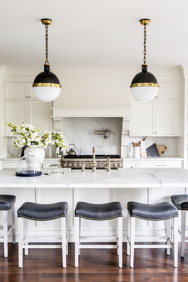 White Kitchen Island with Black Leather Saddle Counter Stools. White Kitchen Island with Black Leather Saddle Counter Stools. Gorgeous kitchen features white shaker cabinets paired with white marble countertops with gray veining and matching backsplash. A white French kitchen hood with corbels stands over a swing arm pot filler and a stainless steel stove. A pair of Hicks Pendants illuminate a long white kitchen island fitted with a farmhouse sink and deck mount vintage faucet lined with black leather saddle seat counter stools. #WhiteKitchenIsland #BlackLeatherSaddleCounterStools white-kitchen-island-with-black-leather-saddle-counter-stools