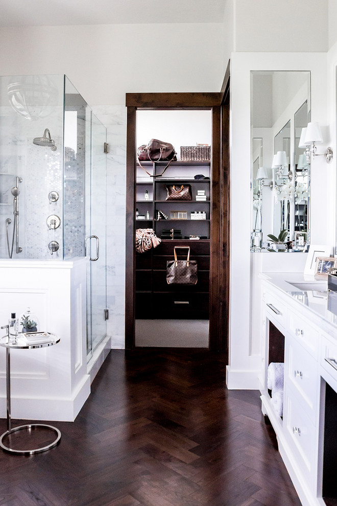 White master bathroom with dark wood trim and dark stained wood herringbone floor. A white master bathroom with dark wood trim leads to a walk-in closet filled with a dark stained closet island and shelves lined with Louis Vuitton bags. Chic master bathroom boasts a glass and wainscoted shower enclosure filled with gray mosaic marble tiles alongside a dark stained wood herringbone floor. wood-herringbone-floor-tile LIV Design Collective.