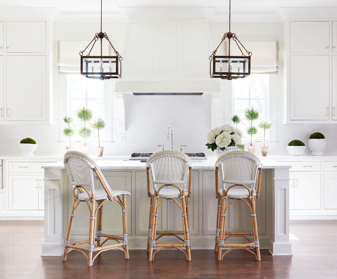  White Kitchen with Black and White French Bistro Counter Stools. All white kitchen features a pair of iron and seeded glass lanterns illuminating a center island topped with honed white marble fitted with a farm sink lined with white and black bistro French counter stools, Serena & Lily Riviera Stools. A white French kitchen hood accented with corbels stands over a white subway tile backsplash and a stainless steel stove. #WhiteKitchen #BlackandWhite #FrenchBistroCounterStools Sarah Bartholomew Design