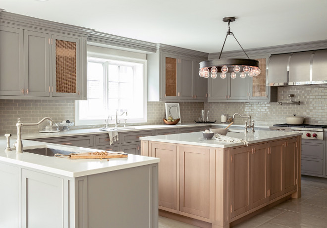 Grey kitchen. Grey kitchen features gray shaker cabinets paired with white quartz countertops and a gray brick tile backsplash. A kitchen peninsula is fitted with a stainless steel sink and satin nickel hook and spout faucet. A secondary kitchen sink stands across from the island and under a window flanked by mesh metal cabinets. A Ralph Lauren Roark Modular Chandelier illuminates a honey colored kitchen island fitted with a prep sink. A stainless steel barrel kitchen hood stands over a swing arm pot filler and an integrated Wolf cooktop accented with pot and pan drawers below. #Greykitchen #shakercabinets #kitchen grey-kitchen RC Studio