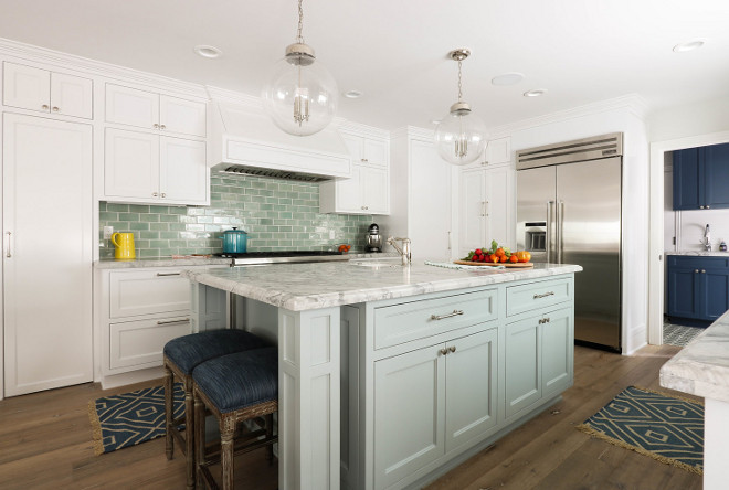 Seafoam kitchen island. White and green kitchen features Regina Andrew Globe Pendants hung over a light gray island framed by gray wash floors and topped with a marble countertop finished with a prep sink and seating denim backless stools. To one side of the island, white industrial sconces are fixed on green subway tiles above window dressed in a bamboo roman shade positioned above a farmhouse sink with a polished nickel faucet mounted in a marble countertop placed a top white inset cabinets finished with polished nickel pulls as upper glass front cabinets are lit by custom lighting. To the other side, a white hood is located above a stainless steel oven range while a double door stainless steel fridge is inset beside white cabinets. sea-foam-kitchen-island Erin Hedrick