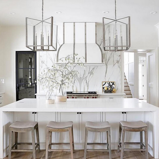  White Enamel Range Hood with Stainless Steel Straps. This all white kitchen features gray saddle counter stools accented with nailhead trim, positioned in front of a white center island finished with a white quartz waterfall countertop illuminated by steel and glass lanterns. Behind the island, a white enamel hood with steel straps is mounted to a gray and white marble slab backsplash above a Wolf range flanked by white cabinets. # WhiteEnamelRangeHood #StainlessSteelStrapsHood Redo Home and Design