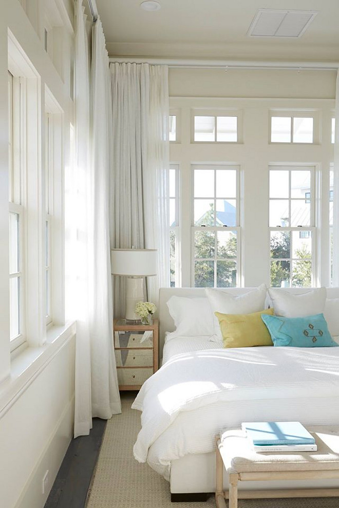  Beach Style Bedroom. This coastal bedroom is decorated with a white upholstered bed dressed in green and blue velvet pillows and a small mirrored nightstand. Wall of windows are dressed in white curtains. cottage-bedroom Geoff Chick & Associates