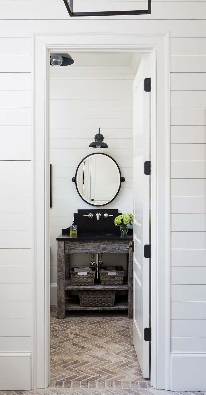 Farmhouse powder room. The designer decided to keep this powder room informal because of its close proximity to the pool. They used weathered wood for the console and leathered black pearl granite for the countertop and backsplash. Light fixture is RH. The brick in the herringbone pattern adds interest to the floor. #farmhouse #powderroom #farmhouseinteriors #shiplap Interiors by Courtney Dickey. Architecture by T.S. Adam Studio.