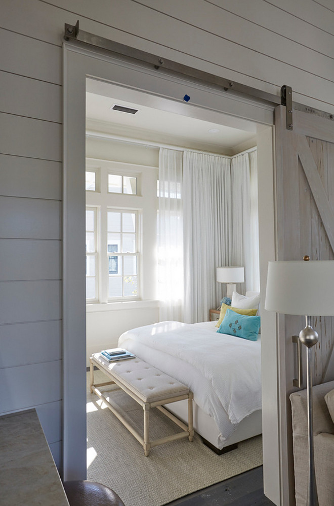 Sliding barn door. Small Bedroom with Pecky Cypress Barn Door on Rails. A pecky cypress barn door on rails opens to a beach style bedroom filled with an off white upholstered bed dressed in green and blue velvet pillows as well as a gray French bench placed at the foot of the bed. sliding-barn-door Geoff Chick & Associates