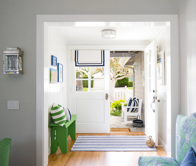 Foyer with Apple Green Bench and Blue Striped Rug. White glass paneled double doors dressed in a blue grosgrain roman shade open to a green, blue, and white beach cottage foyer featuring an apple green bench topped with a green awning stripe pillow positioned behind a blue striped rug. Lighting is an octagon flush mount. Grey paint color is "Frazee Foggy London". Foyer with Apple Green Bench and Blue Striped Rug #Foyer #AppleGreenDecor #Bench #BlueStripedRug