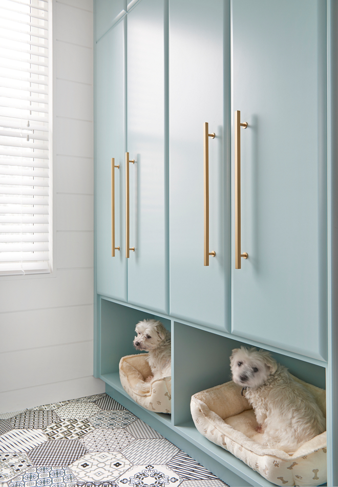 White and black mosaic floor tiles lead to stacked tall turquoise laundry room cabinets in Gossamer Blue by Benjamin Moore accented with brass pulls and fixed above built in dog beds. Flooring is Ceragres St-Barth 8 x 8 hex with laticrete dark grey grout. Walls are finished with a white shiplap trim positioned framing a window. #turquoiselaundryroom #cabinets #GossamerBluebyBenjaminMoore #brasspulls #builtindogbeds Soda Pop Design Inc.