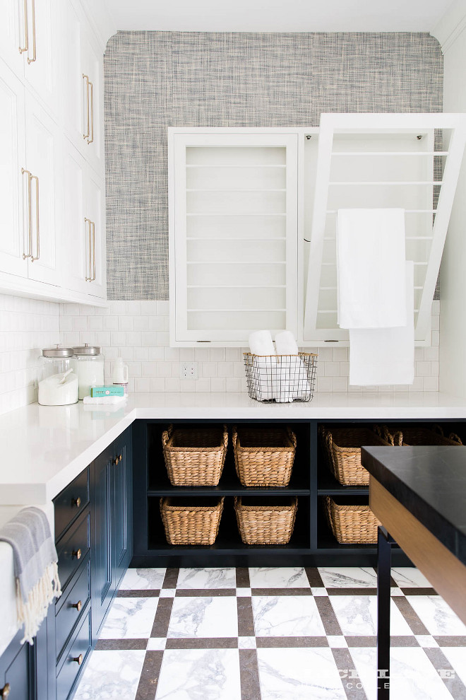 Laundry Room Drying Racks. Laundry room Side by Side Drop Down Drying Racks. White and gray marble floor tiles complement dark blue cabinets contrasted with a white quartz quartz countertop fixed against white porcelain square stacked wall tiles and beneath white stacked upper cabinets finished with satin nickel pulls. Under the countertop on adjacent wall, dark blue shelves are filled with wicker baskets and located under two white side-by-side pull down drying racks partially fixed to gray grasscloth wallpaper #laundryroom #DryingRacks