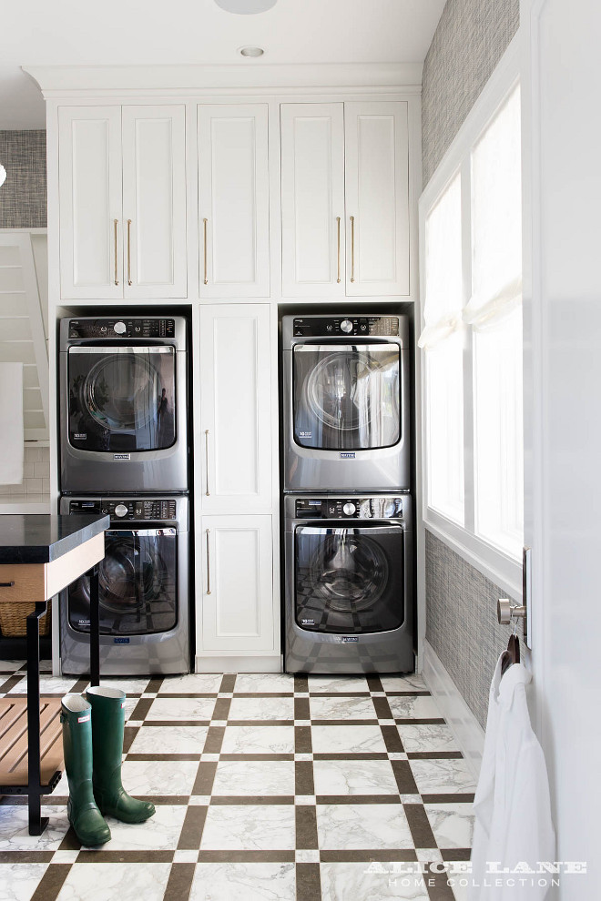 Laundry Room. Laundry room with Pull Out Cabinets Between Stacked Washers and Dryers. This laundry room is equipped with two sets of stacked Maytag washers and dryers placed on gray and white marble floor tiles enclosed on either side of and beneath white cabinets adorning long brass pulls. An adjacent wall features windows dressed in white roman shades and framed with gray grasscloth wallpaper. #LaundryRoom