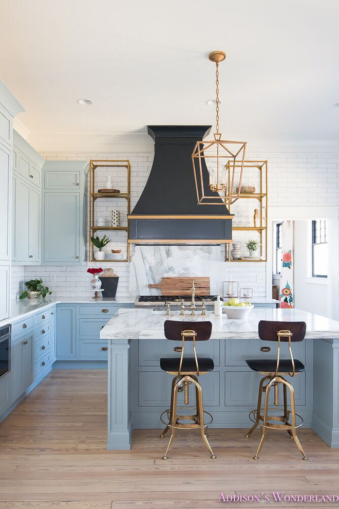 Blue Kitchen with Brass accents. A black French kitchen hood, painted in Sherwin Williams Inkwell, is accented with gold trim and it's flanked by brass shelving. Blue kitchen features blue shaker cabinets painted in a custom color, 50% of Sherwin Williams Stardew and 50% Uncertain Gray, paired with Calcutta Reale Marble countertops and a linear white tile backsplash. A black French kitchen hood painted Sherwin Williams Inkwell is accented with gold trim, flanked by brass shelving units mounted on the wall stands over a gray marble cooktop backsplash, Calcutta Reale Marble. A Darlana Medium Lantern illuminates a blue kitchen island fitted with a marble apron sink as well as a satin nickel vintage faucet lined with gold and black vintage architect's stools, Restoration Hardware Vintage Toledo Bar Chairs. Blue Kitchen with Brass accents. Blue Kitchen with Brass accents. #BlueKitchen #Brassaccents #kitchenbrass Home Bunch's Beautiful Homes of Instagram @addisonswonderland