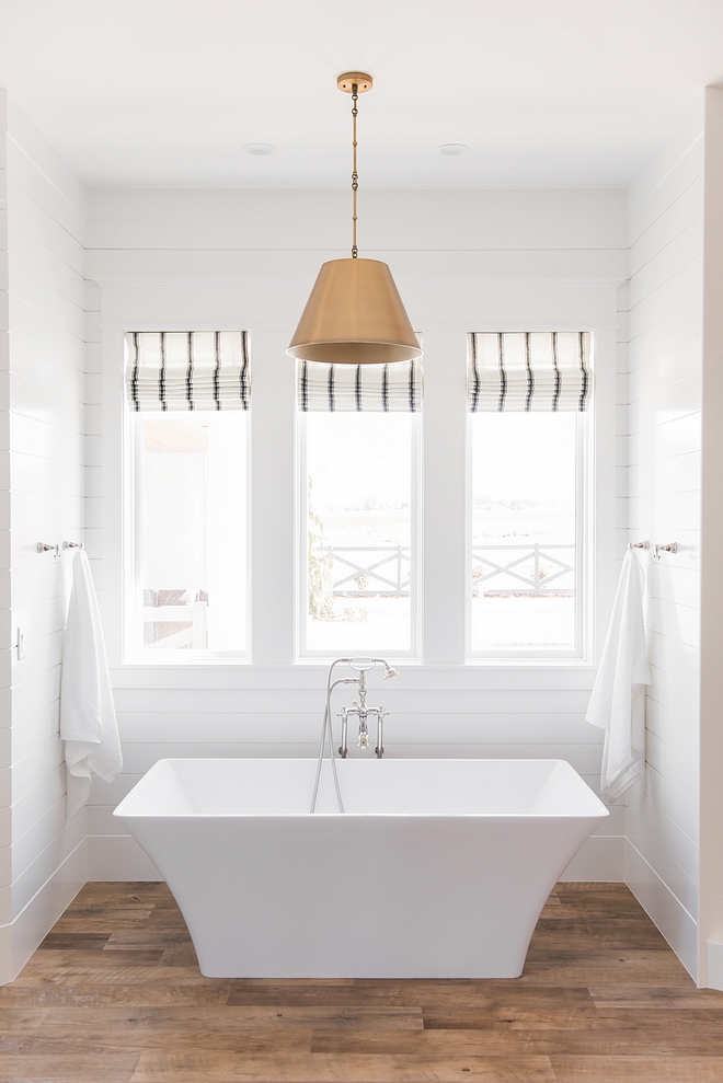 Modern farmhouse bathroom with shiplap freestanding tub, plank vinyl flooring, brass cone pendant and striped back and white roman shades