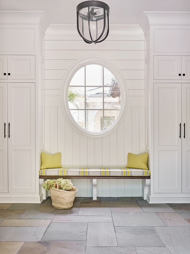 A custom mudroom bench is flanked by closed lockers and it features an oval window surrounded by a mix of vertical and horizontal shiplap. Cabinets are are custom inset cabinets with a fixed panel and crown moulding above. Paint color is White Dove OC-17 Benjamin Moore #mudroom #ovalwindow #mudrooms #shiplap #verticalshiplap #horizontalshiplap #mudroombench #cabinet