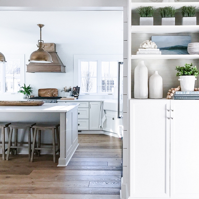 Coastal farmhouse kitchen The living room opens to a large and bright white kitchen with hardwood flooring Notice the beautifully styled bookcase on the right #Coastalfarmhousekitchen #Coastalfarmhouse #kitchen #hardwoodfloors #bookcase