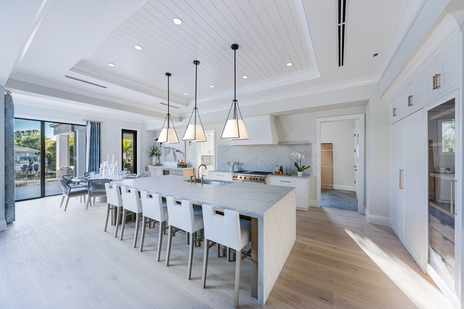 The expansive white kitchen showcases custom white cabinets grand center island finished in White Oak and a comfortable dining space with water views and a gorgeous butlers pantry #whitekitchen #whitecabinets #island #WhiteOak #diningspace #butlerspantry