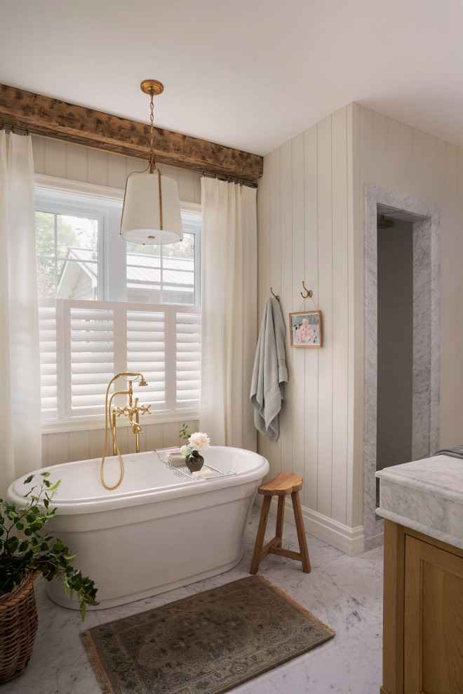 French country primary bathroom with shiplap walls painted Benjamin Moore Seapearl, freestanding tub, Carrara marble floor, and Antique Gild pendant.