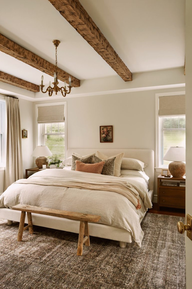 Primary French country bedroom with Benjamin Moore Seapearl walls, neutral bedding, rustic ceiling beams, and vintage wood bench.