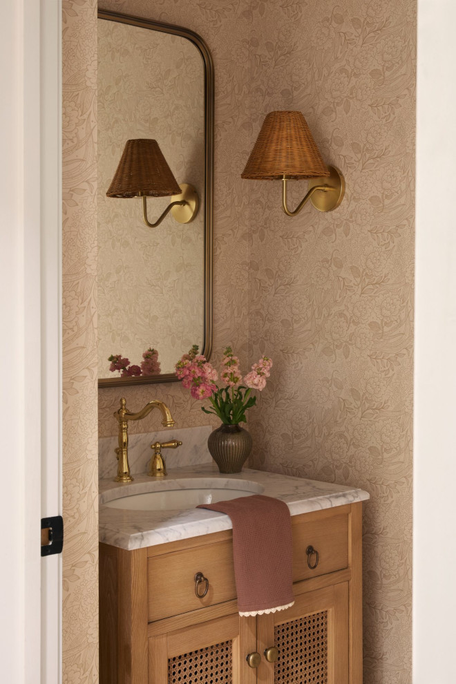 Earth-Tone Powder Room Vanity with Natural Wood and Rattan Sconce Warm earth-tone powder room featuring a natural wood vanity with cane doors, brass faucet, rattan wall sconce, marble countertop, and soft floral wallpaper.