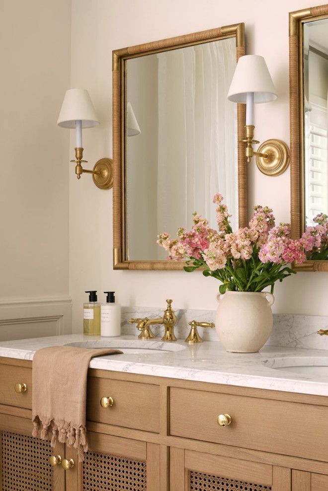 Natural Wood Double Vanity with Brass Fixtures Neutral bathroom featuring a natural wood double vanity, brass fixtures, marble countertop, white sconces, and soft pink floral decor.