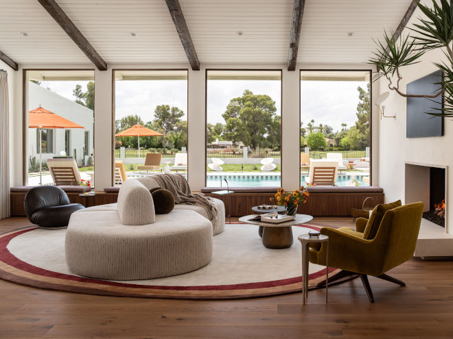 Mid-century modern living room with sloped ceiling, wall-to-wall window seat, and open views of backyard pool.