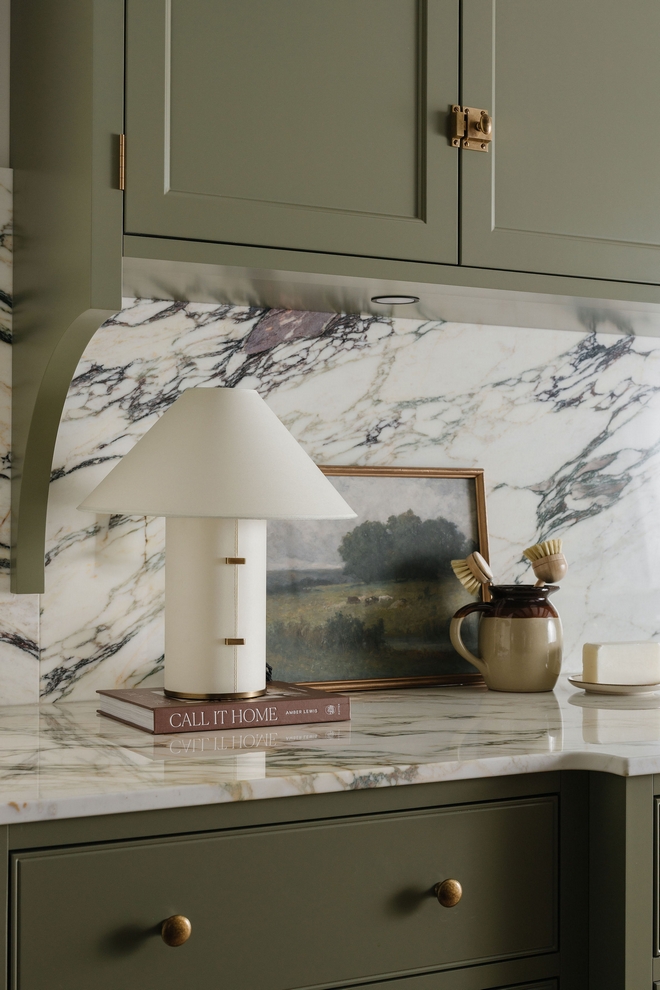 Close-up of Calacatta Viola marble backsplash with kitchen styling including a table lamp, framed artwork, and neutral decor on a marble countertop.