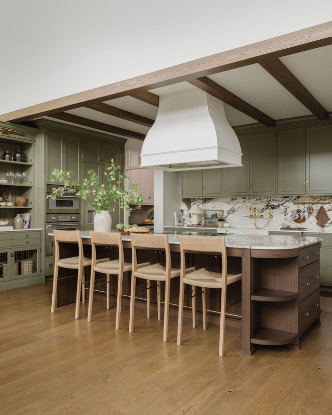 Open-concept classic kitchen with green cabinets, wood island, marble countertops, bar stools, exposed beams, and white range hood.