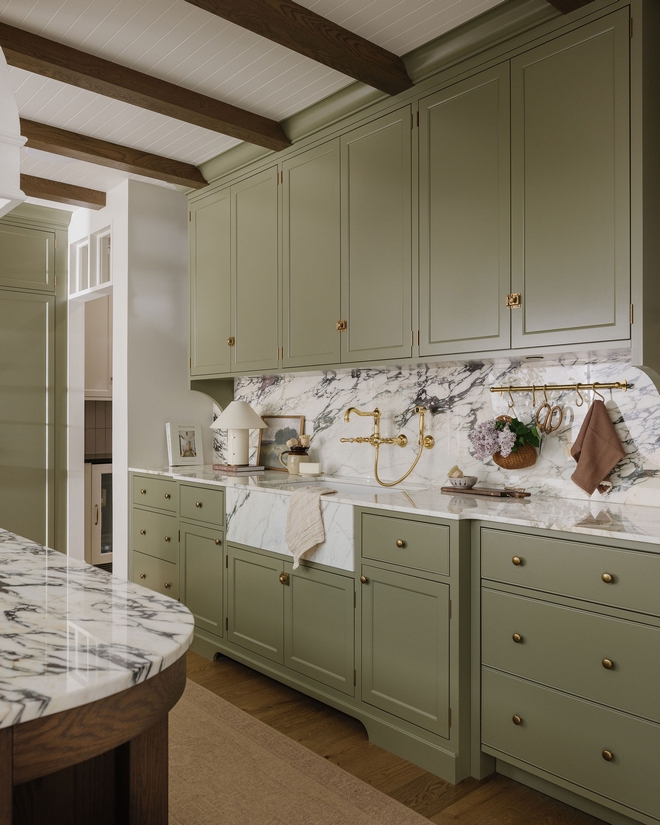 Wide view of kitchen with green cabinetry, Calacatta Viola marble slab backsplash, brass rail with hanging utensils, and integrated sink area.
