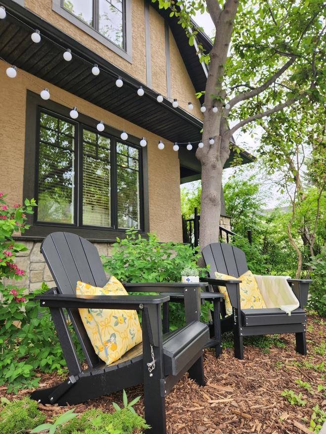 White Adirondack chairs styled with yellow outdoor pillows, fresh flowers, and a matching side table on a sunny front yard patio.