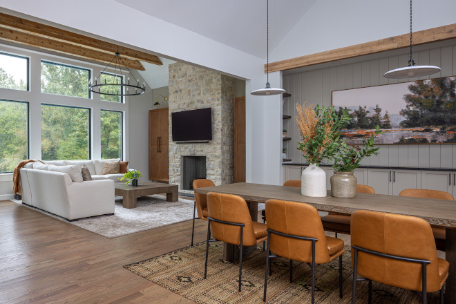open-concept-living-room-stone-fireplace Open-concept living room and dining room featuring a floor-to-ceiling stone fireplace, vaulted ceilings, exposed wood beams, large windows, and warm leather dining chairs.