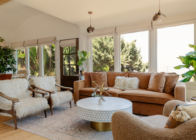 Sunlit living room with a fawn velvet sofa, vintage sheepskin chairs, a blue-and-white inlay coffee table, and cream Roman shades.