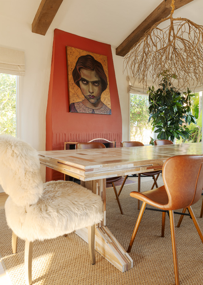 Reclaimed wood dining table with sheepskin and brown chairs beneath a branch chandelier, facing a coral-red fireplace chimney with a large portrait.