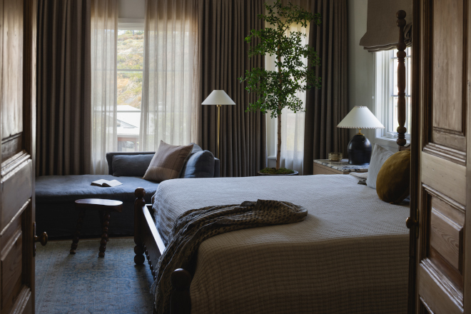 Layered-Modern-English-Country-Bedroom View through antique wood doors into a layered English Country bedroom with dark wood bed, taupe linens and charcoal chaise.