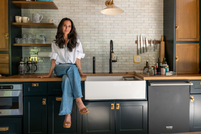 Alycia Lane Joseph seated on an oak countertop beside a white farmhouse sink, with Hague Blue cabinets, brass hardware, and white Zellige tile.