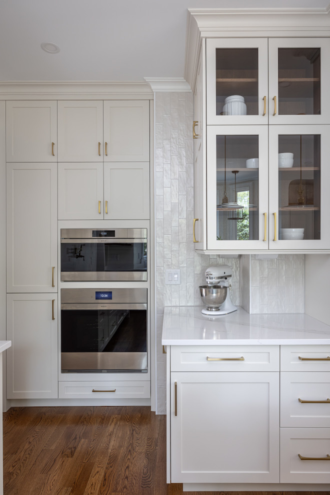 Custom White Kitchen Cabinets with Built-In Wolf Ovens and Glass Cabinets Floor-to-ceiling muted taupe kitchen cabinetry with two stacked stainless steel Wolf ovens, brass cabinet pulls, glass-front display cabinets, and a dedicated stand mixer countertop.