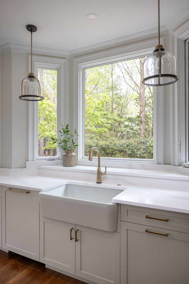White Farmhouse Kitchen Sink with Brass Faucet and Large Windows White apron-front farmhouse sink beneath a large picture window, framed by smaller windows, with a brushed brass faucet, white quartz countertops, and glass pendant lights.