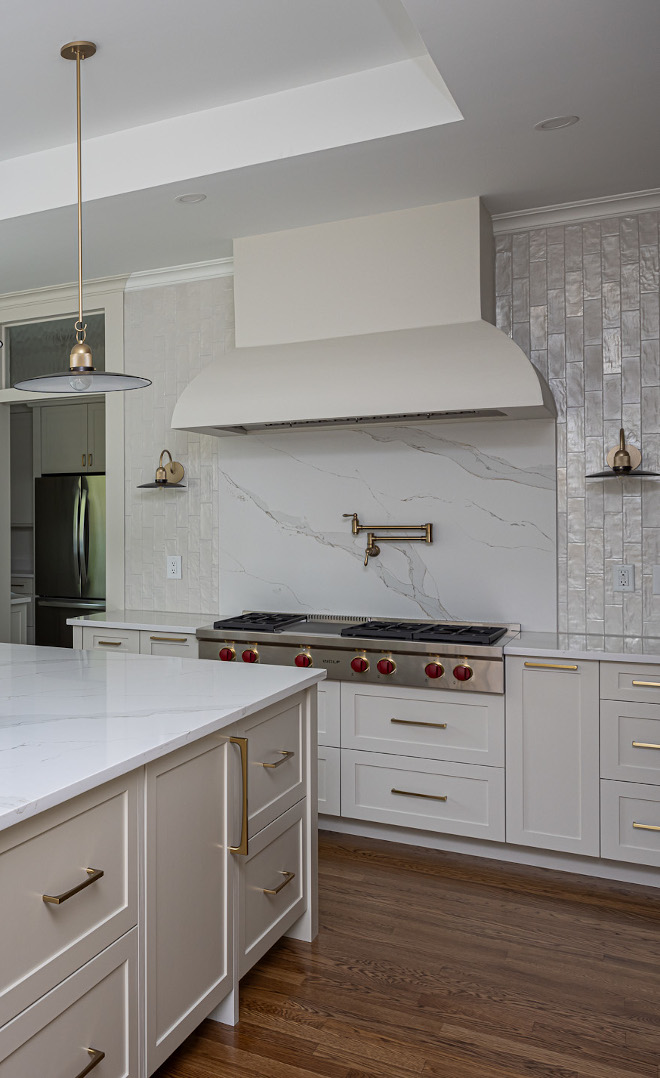 White Kitchen with Custom Range Hood, Quartz Backsplash and Brass Pot Filler Bright white kitchen featuring a custom plaster-look range hood, quartz slab backsplash, brass pot filler, large center island, and warm hardwood flooring.