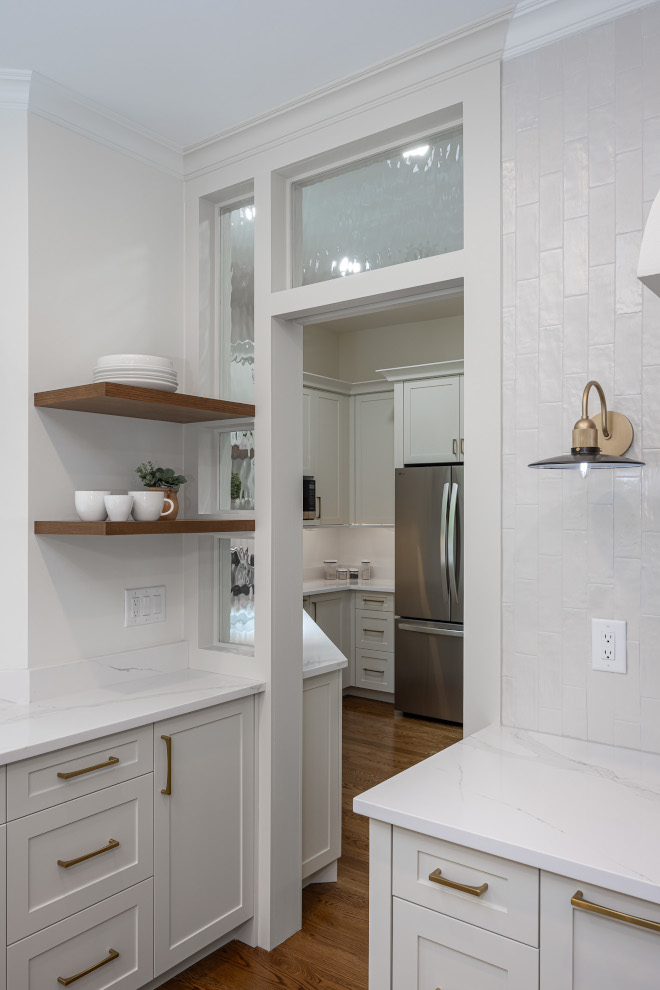White Kitchen with Floating Wood Shelves and Textured Tile Backsplash White kitchen with natural wood floating shelves, brass wall sconce, textured white ceramic tile, quartz countertops, and passageway to a secondary prep kitchen.