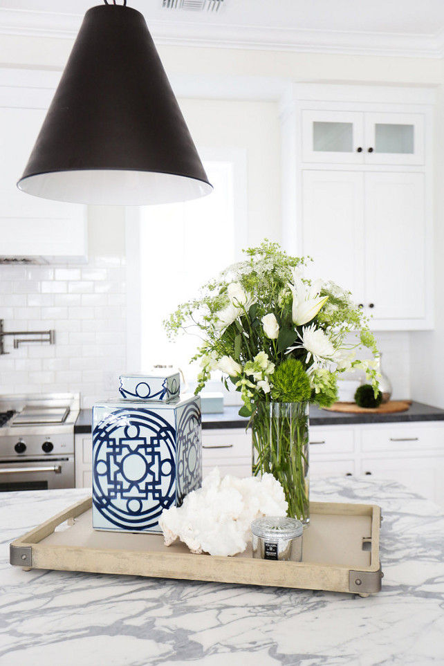 Kitchen Ideas. Stunning kitchen features a Goodman Hanging Lamp illuminating a grey and white marble top island and a wood tray with metal brackets. A white kitchen hood, flanked by windows, stands over a white brick tiled backsplash and a stainless steel stove. The countertop is Carrara Marble. #Kitchen #KitchenIdeas