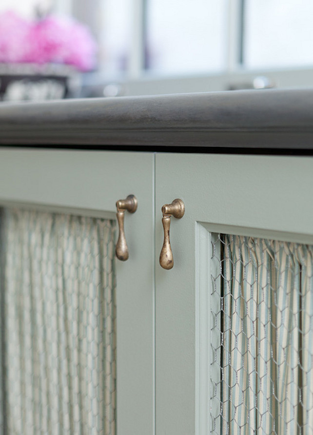 Chicken Wire Cabinet Door. This country kitchen boasts gray cabinets accented with chicken wire doors lined with striped fabric with antique brass hardware under honed black countertops. #ChickenWire #Cabinet #Kitchen Jenny Wolf Interiors.