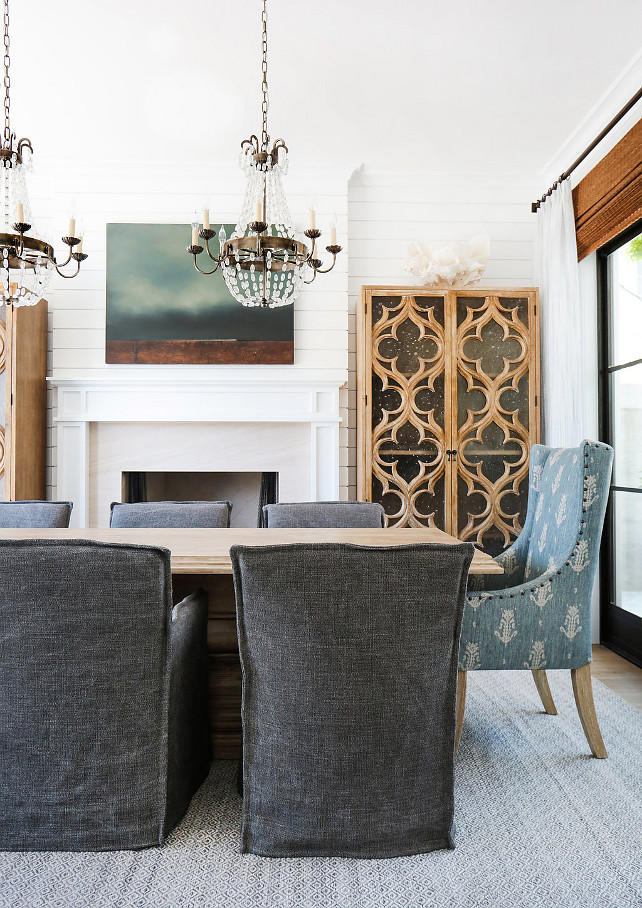 Dining Room Design. This gorgeous dining room features a pair of Paris flea market chandeliers over a wood dining table lined with charcoal gray slipcovered dining chairs as well as blue wingback dining chairs. You will also notice the shiplap accent wall and fireplace flanked by quatrefoil glass cabinets. The glass and steel doors are dressed in bamboo roman shades and white linen draperies. #DiningRoom