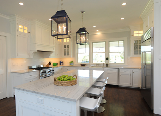 Kitchen Cottage. Cottage Kitchen Design. White Cottage Kitchen. White coastal kitchen Ideas. Stunning U-shaped kitchen with white cabinetry accented with brushed nickel hardware alongside white quartzite countertops with a subway tiled backsplash. The kitchen features a stainless steel stove below a white paneled stove hood accented with corbels across from a stainless steel industrial style, built-in refrigerator. A farm sink stands below three sash windows opposite an extra long kitchen island lined with Piston Barstools illuminated by a pair of lacquered navy lantern pendants from Oomph. Nina Liddle Design.