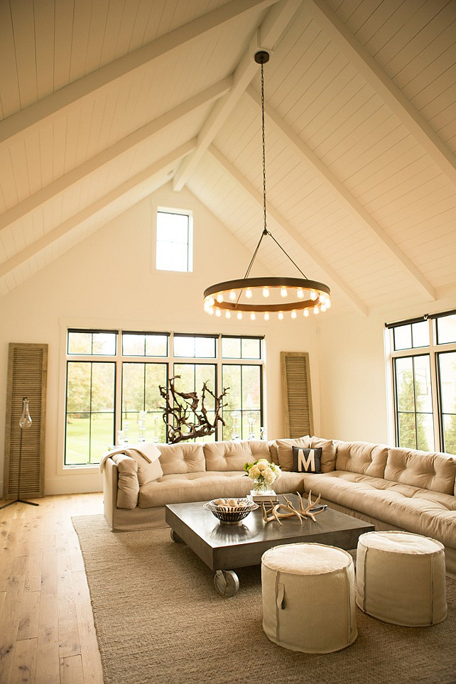 Industrial Living Room. Industrial living room features a paneled cathedral ceiling accented with a Ralph Lauren Roark Modular Chandelier illuminating a beige tufted slipcovered sectional facing a metal industrial coffee table on wheels and a pair of round poufs atop a jute rug. #LivingRoom #Industrial #IndustrialInteriors Hahn Builders.