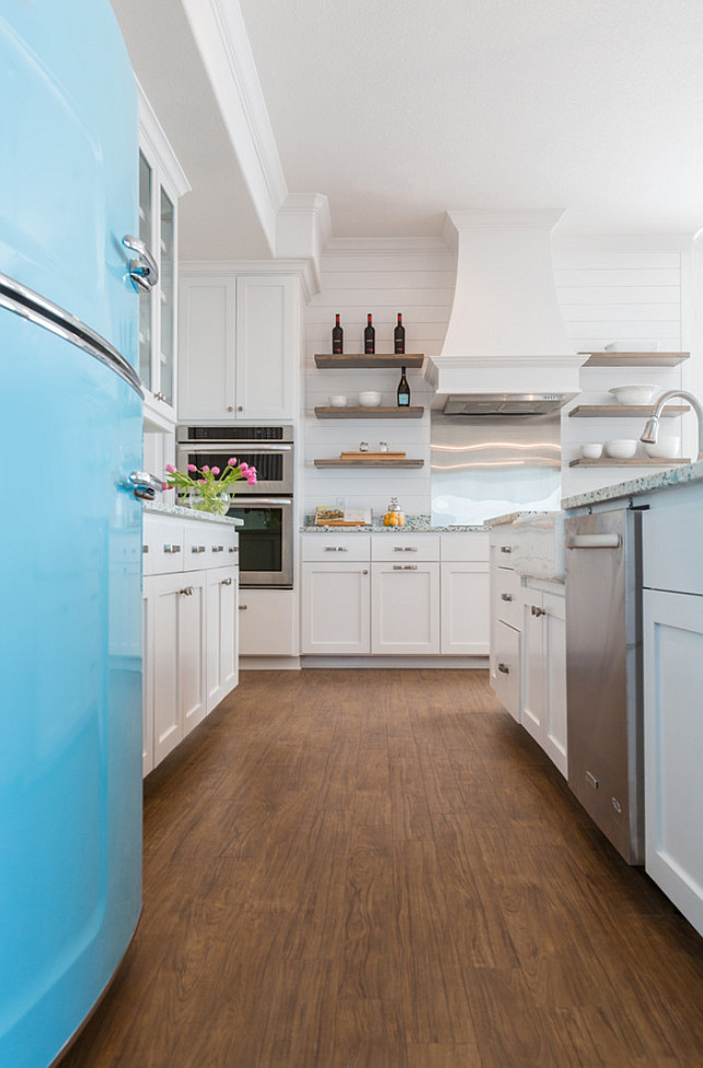White and turquoise cottage kitchen features an extra-long island fitted with recycled glass countertops framing sink and satin nickel gooseneck faucet. A turquoise refrigerator by Big Chill stands beside glass-front cabinets across from a white kitchen hood which is flanked by stacked floating shelves situated over an induction cooktop and a stainless steel cooktop backsplash next to microwave and oven.