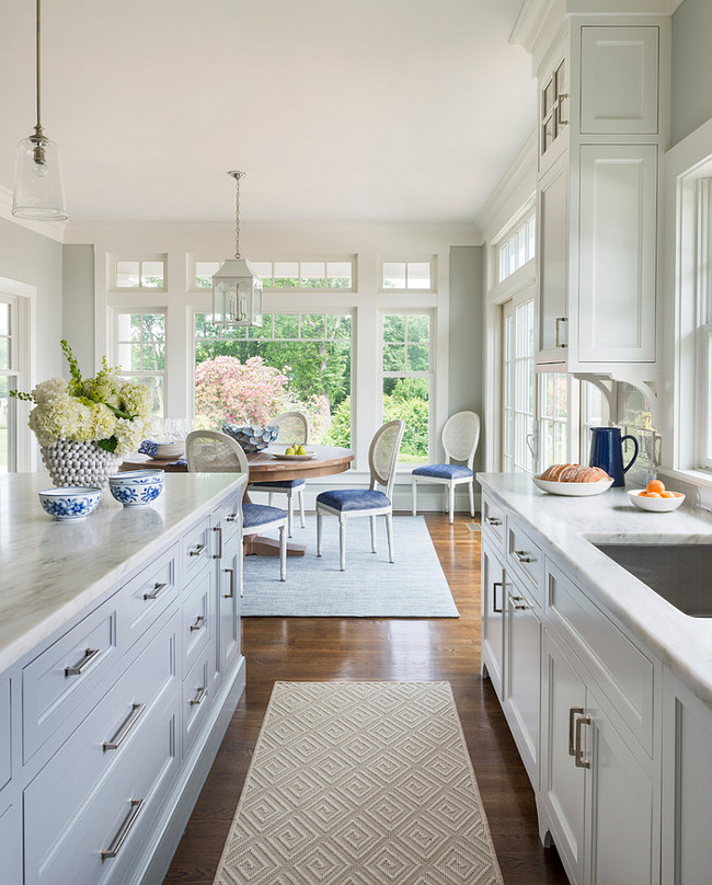 White and gray kitchen with navy blue accents. White and gray kitchen features white cabinets paired with white marble countertops fitted with a stainless steel sink. Clear glass pendants hang over a gray kitchen island topped with white marble alongside a gray geometric runner and navy blue accents. Digs Design Company. 