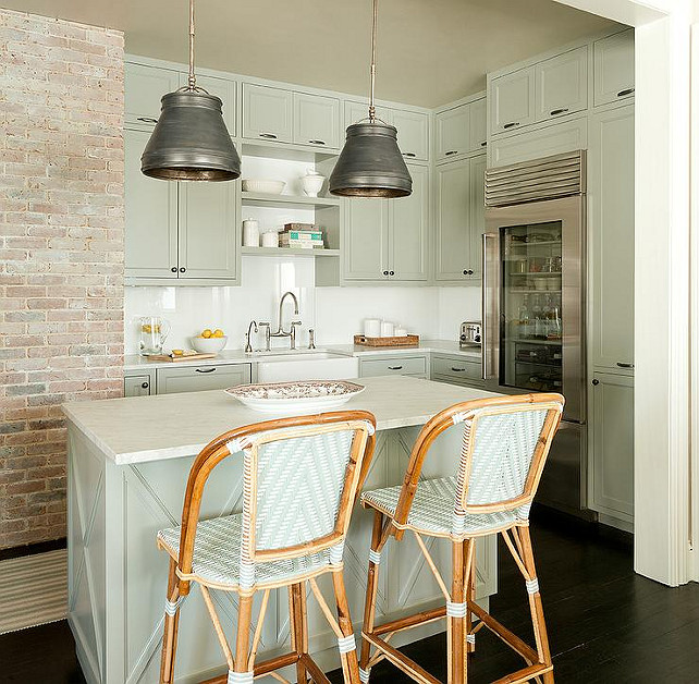 Small Kitchen. Small kitchen features a pair of metal pendants illuminating a gray green center island lined with green French bistro counter stools. Kitchen with gray green cabinets paired with honed white marble countertops and a white quartz tiled backsplash. Kitchen boasts stacked shelving over a small farmhouse sink and a deck mount faucet next to a glass front refrigerator. Jenny Wolf Interiors.