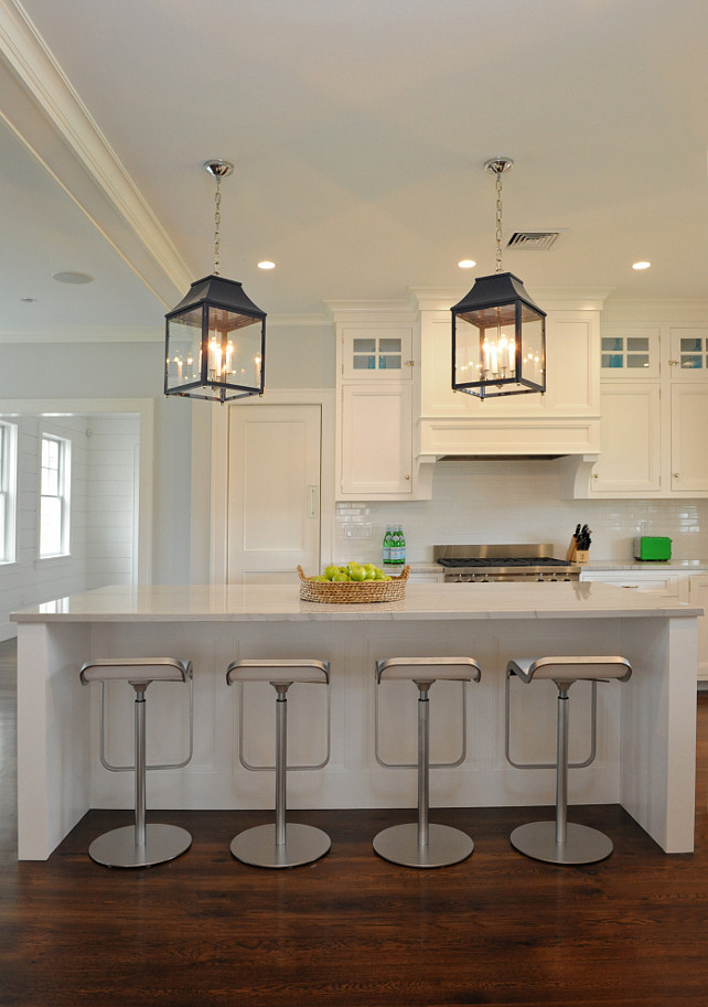 White Kitchen. Coastal White Kitchen. Modern Coastal White Kitchen. Transitional Coastal White Kitchen. Kitchen with white, flush front cabinets accented with brushed nickel pulls and white quartzite counters which highlight a stainless steel stove with subway tiled backsplash and paneled stove hood accented with corbels. The kitchen features an elongated kitchen island with Piston Barstools tucked below a white quartzite countertop lit by a pair of lacquered navy lantern pendants. The designer used a pair of the Oomph Palo Alto Lantern in this kitchen. Nina Liddle Design.