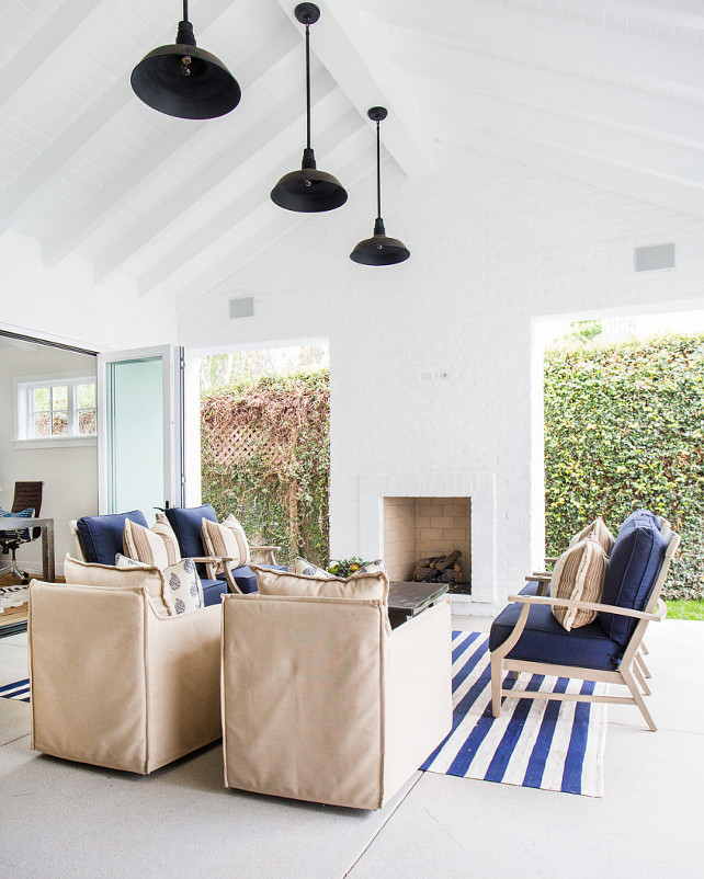 This covered patio with vaulted ceiling features white wood beams and black vintage barn pendants. Natural linen chairs faces an outdoor white brick fireplace flanked by open doorways across from a reclaimed wood coffee table and blue chairs. A blue striped rug adds a coastal feel to the space.