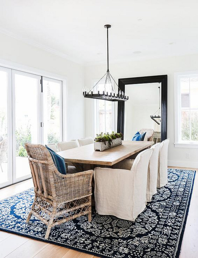 Dining room features an iron linear chandelier illuminating a reclaimed wood trestle dining table lined with natural linen slipcovered dining chairs as well as wicker chairs placed at each end of the table atop a black and white medallion rug placed in front of a black beveled leaning mirror. #DiningRoom