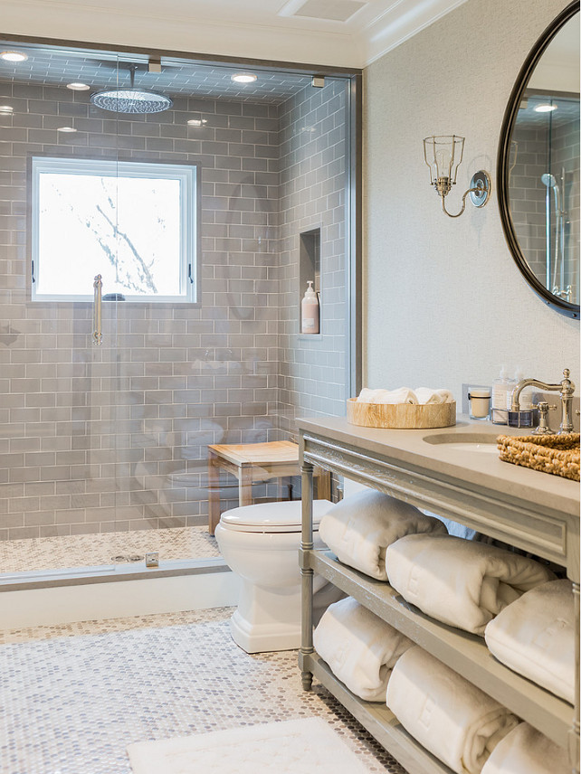 Bathroom. Gray Bathroom. Gray Bathroom Vanity. Gray bathroom features gray walls framing a round rivet mirror illuminated by uplight cage sconce over a gray French bath vanity with shelves filled with fluffy white towels topped with gray countertop framing a round sink paired with a hook and spout faucet beside toilet atop white and gray penny tiled floor. A master bathroom shower boasts gray subway tile surround framing window and tiled niche alongside a ceiling mounted rain shower head over white and gray penny tiled shower floor finished with a seamless glass shower door. #Bathroom #GrayBathroom Brookes and Hill Custom Builders. 