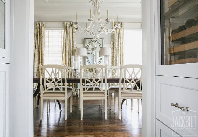 Traditional Dining Room. E. F. Chapman Oslo Chandelier. Traditional dining room features a chippendale dining table lined with white French dining chairs illuminated by an E. F. Chapman Oslo Chandelier. French dining room boasts a French blue sideboard cabinet topped with crystal buffet lamps under a beaded mirror flanked by windows dressed in yellow and gray chintz curtains. #Tradtional #DiningRoom JackBilt Homes.